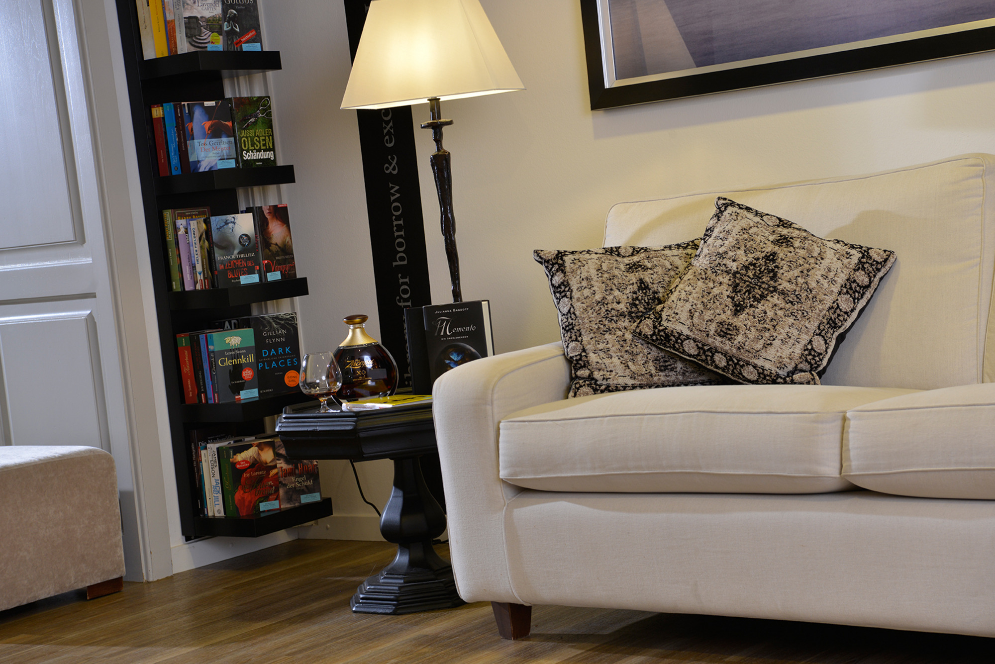 Gemütliche Wohnecke mit weißem Sofa, gemusterten Kissen, schwarzer Stehlampe und Bücherregal vor hellen Wänden und Holzfußboden.