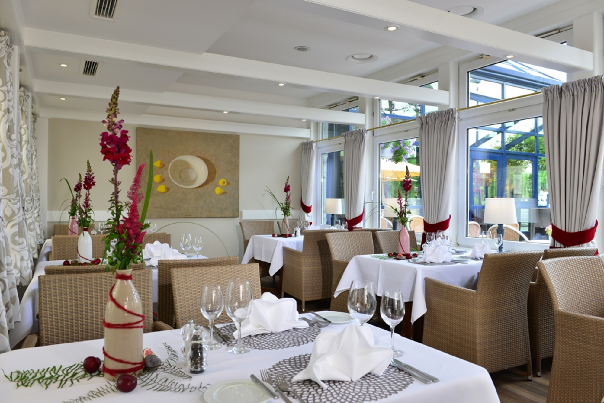 Restaurant mit weiß gedeckten Tischen, rot-weiß gefalteten Servietten, Gläsern und Besteck – lichtdurchflutet durch große Fenster.