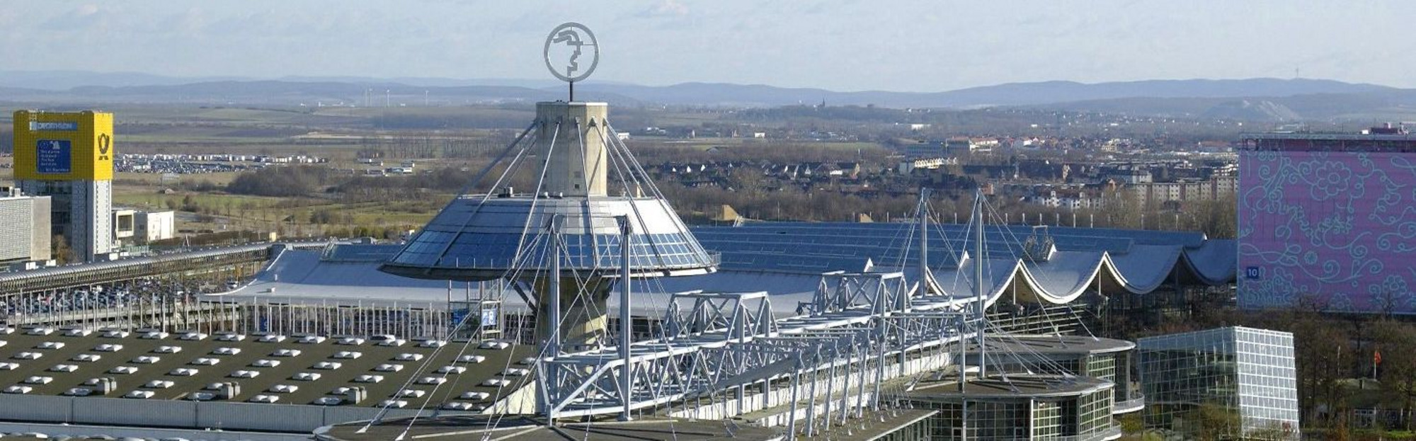Luftaufnahme der Deutschen Messe Hannover mit mehreren Messehallen, markantem Turm mit weißer Kuppel, umgeben von Straßen, Parkplätzen und Grünflächen.
