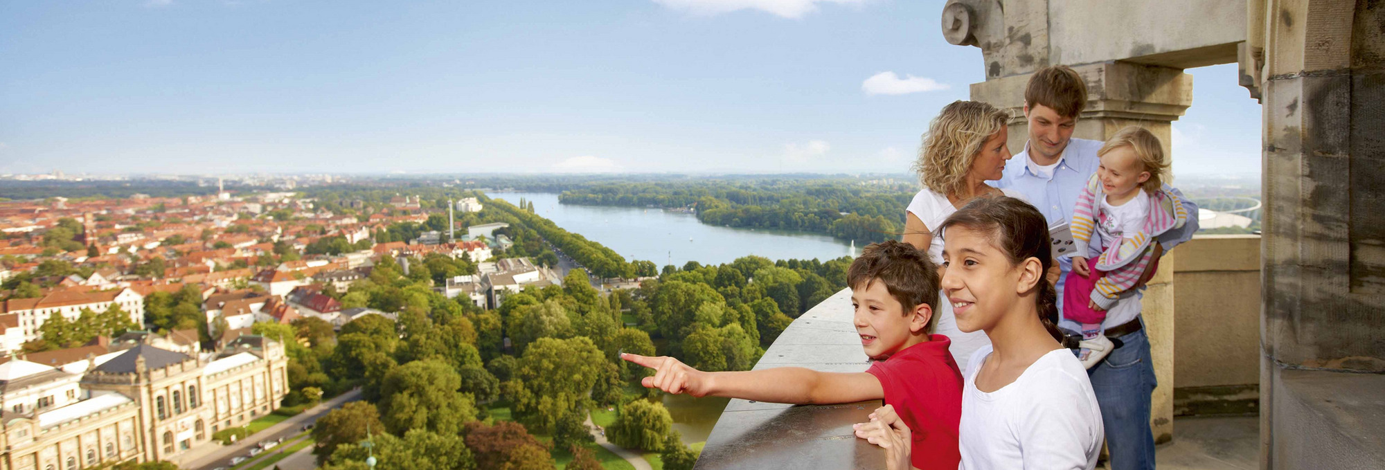 Menschen auf einer Terrasse mit Blick auf die Stadt Hannover, Fluss, Bäume und Gebäude unter klarem Himmel mit wenigen Wolken.