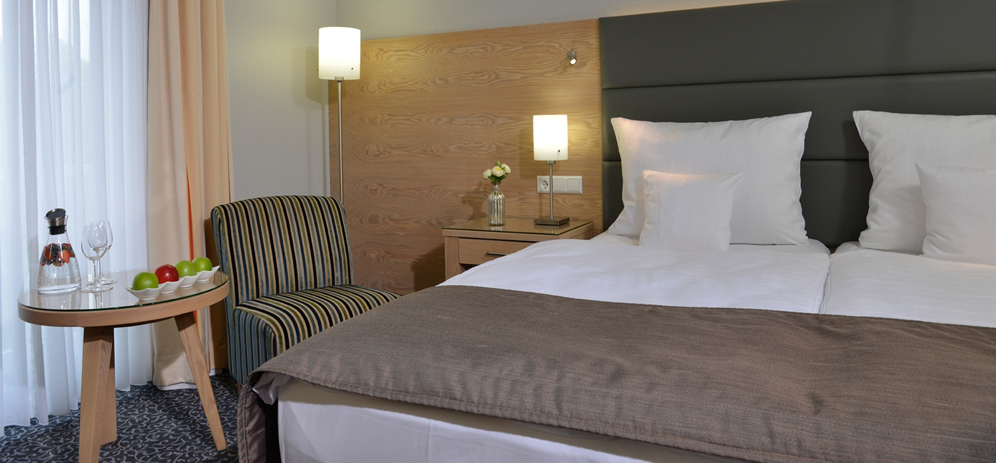Modernes Hotelzimmer mit Doppelbett, Nachttischlampe, Sessel und Tisch mit Obst und Wasserflasche.