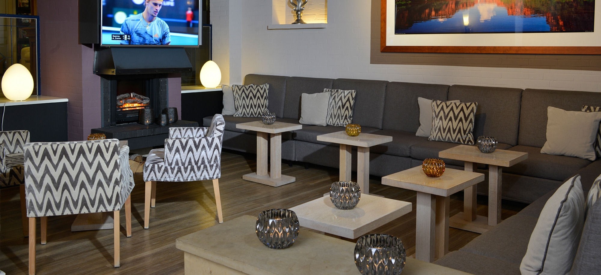 Modernes Kaminzimmer mit Holzfußboden, gemütlichen Sitzgruppen, Wand-TV, Kunstwerken und warmem Licht durch Pendelleuchten.