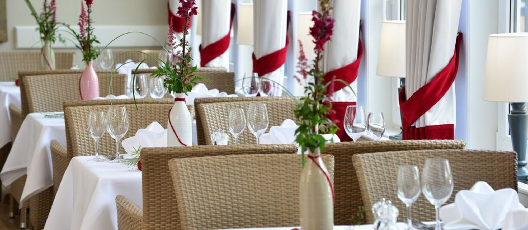 Restaurant mit weiß gedeckten Tischen, rot-weiß gefalteten Servietten, Gläsern und Besteck – lichtdurchflutet durch große Fenster.