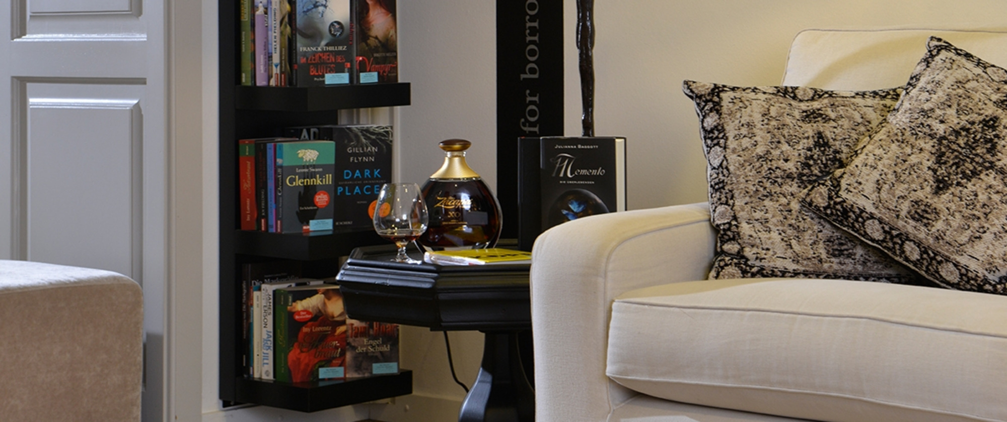 Gemütliche Wohnecke mit weißem Sofa, gemusterten Kissen, schwarzer Stehlampe und Bücherregal vor hellen Wänden und Holzfußboden.