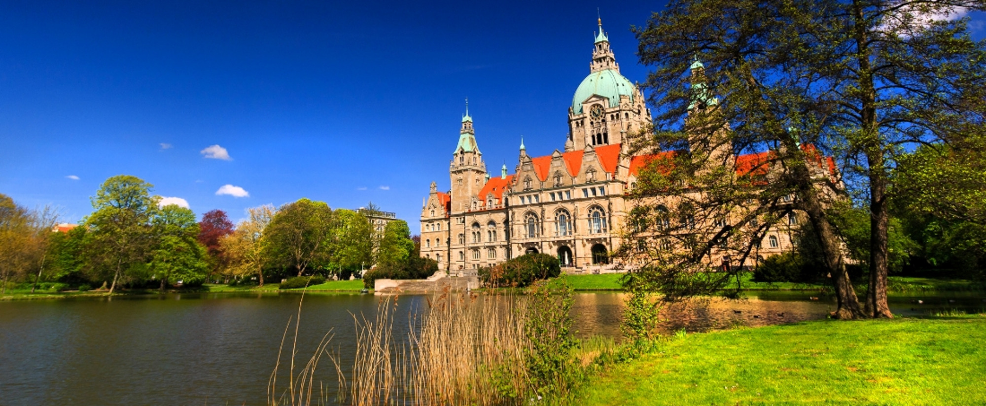 Neues Rathaus Hannover mit großer Kuppel und Türmen, gelegen an einem Teich, umgeben von Bäumen und Grün bei klarem, sonnigem Himmel.