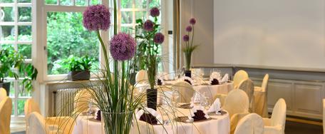 Eleganter Bankettsaal mit runden Tischen, weißen Tischdecken, Blumenschmuck, Stuhlhussen mit Goldschleifen und großen Fenstern.