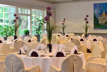 Eleganter Bankettsaal mit runden Tischen, weißen Tischdecken, Blumenschmuck, Stuhlhussen mit Goldschleifen und großen Fenstern.