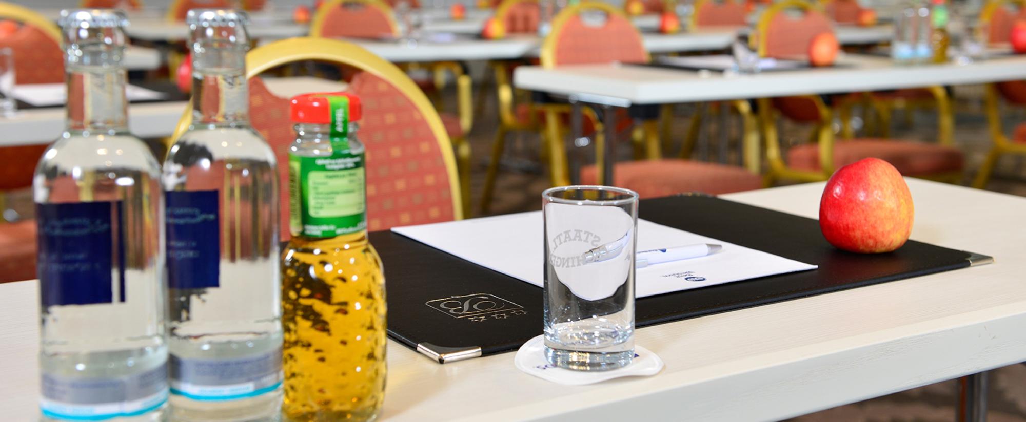 Tagungsraum mit Tischreihen, roten Stühlen, Notizmaterial, Wasser, Säften und Äpfeln auf jedem Platz.