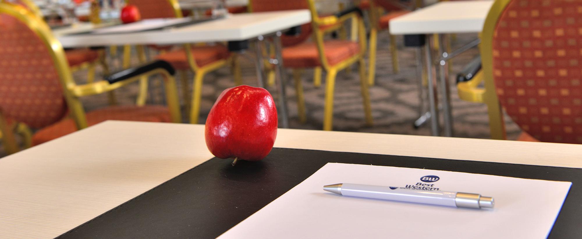 Tagungsraum mit Tischreihen, roten Stühlen, Notizmaterial und Äpfeln auf jedem Platz.