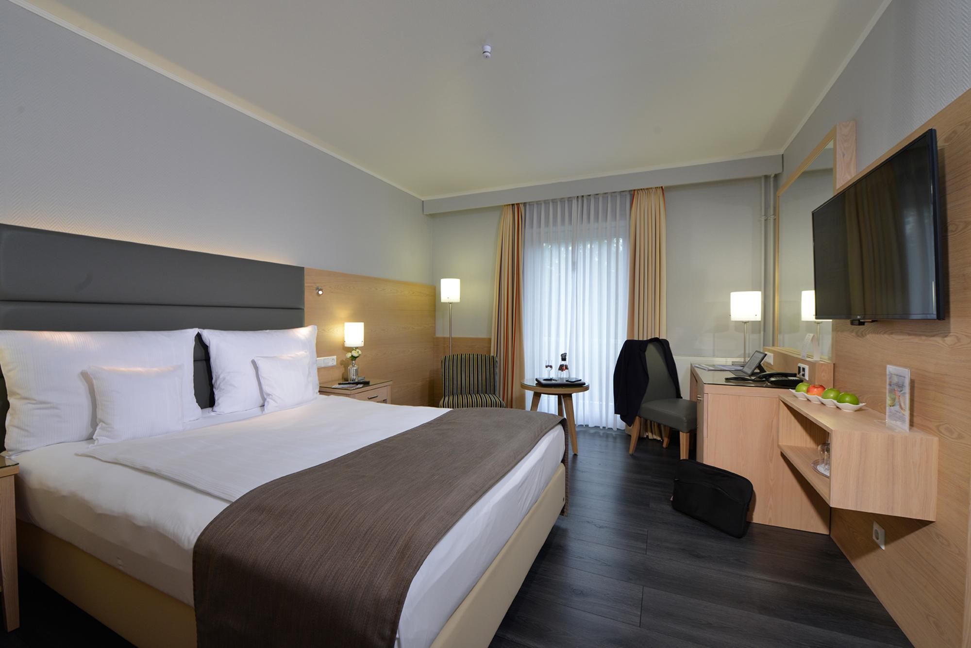 Modernes Hotelzimmer mit großem Bett, weißer Bettwäsche, Flachbild-TV, großen Fenstern und neutraler Deko.