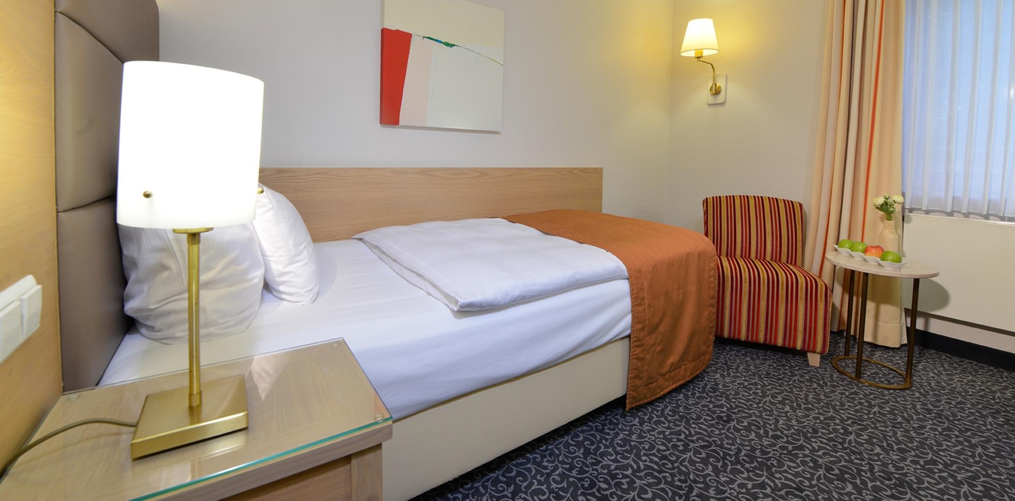 Komfortables Hotelzimmer mit Doppelbett, Nachttischlampe, Sessel, Schreibtisch und heller Beleuchtung.