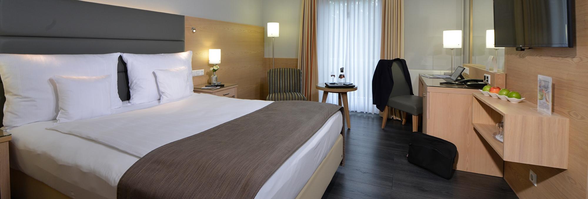 Modern eingerichtetes Hotelzimmer mit großem Doppelbett, Schreibtisch, Sitzbereich, Flachbildfernseher und Tageslicht.