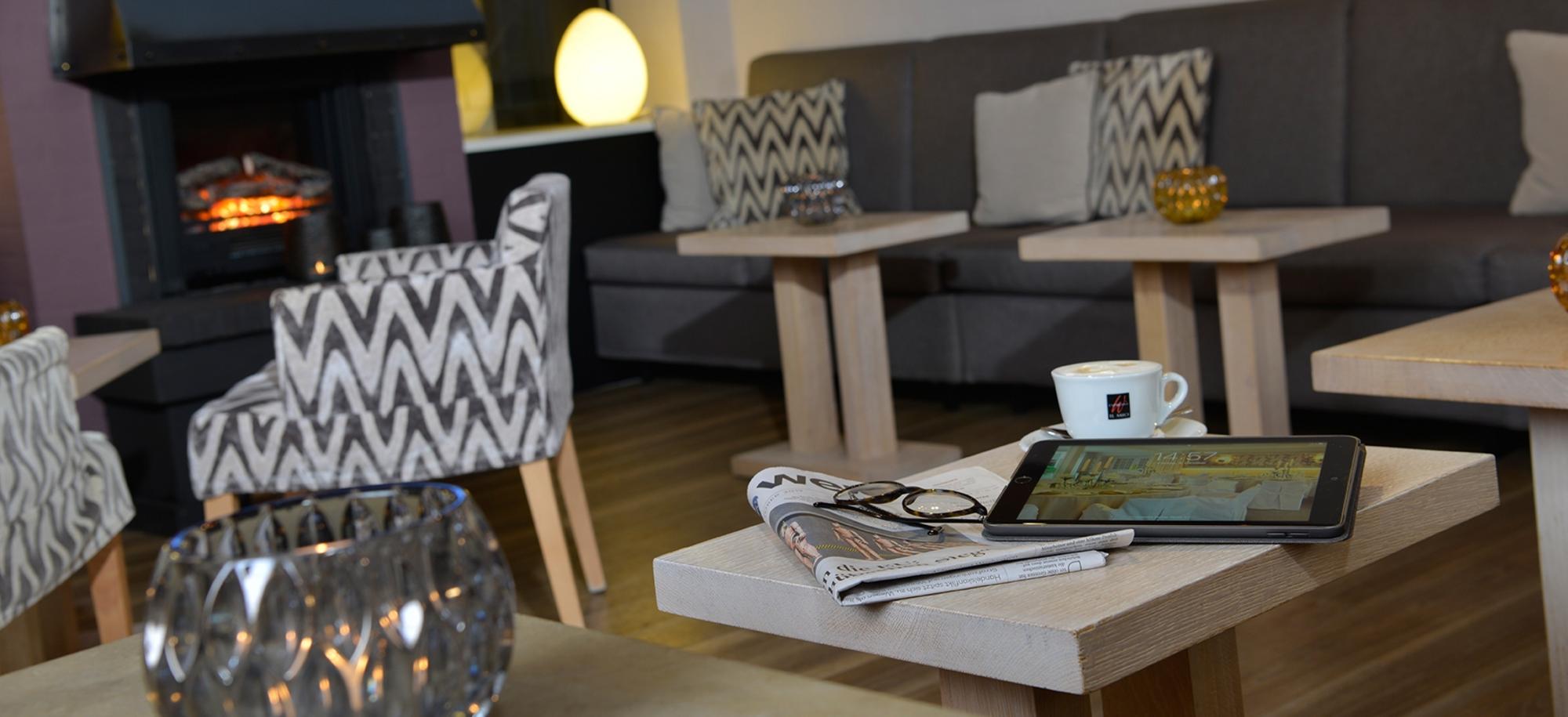 Gemütliches Kaminzimmer mit Kamin, Holztisch, Feuerholzkorb, weißen Sesseln, Sofa, Kissen und warmer Beleuchtung.