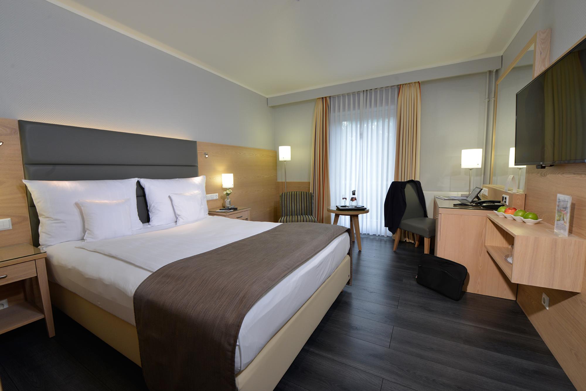 Modernes Hotelzimmer mit großem Bett, weißer Bettwäsche, Flachbild-TV, großen Fenstern und neutraler Deko.