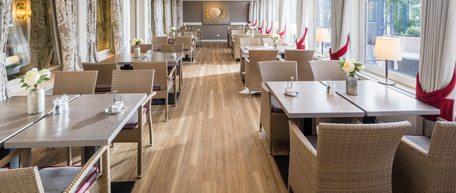 Eleganter Speisesaal mit weiß gedeckten Tischen, Blumenschmuck und viel Tageslicht durch große Fenster.