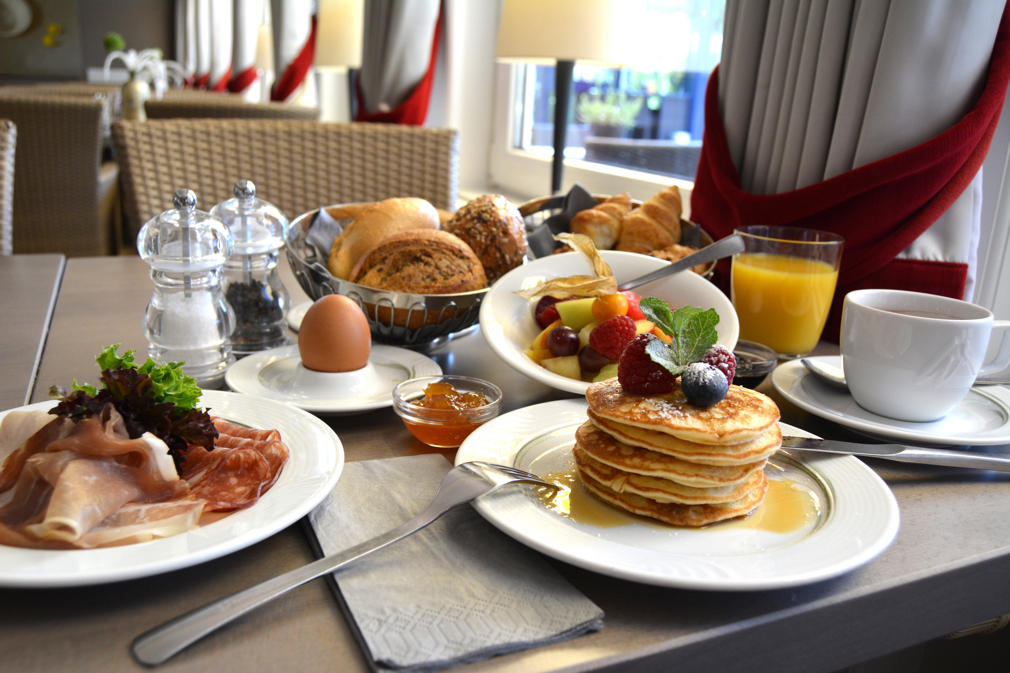 Frühstückstisch mit Pancakes, Eiern, Speck, Obstsalat, Saft und Kaffee – einladend und reich gedeckt.