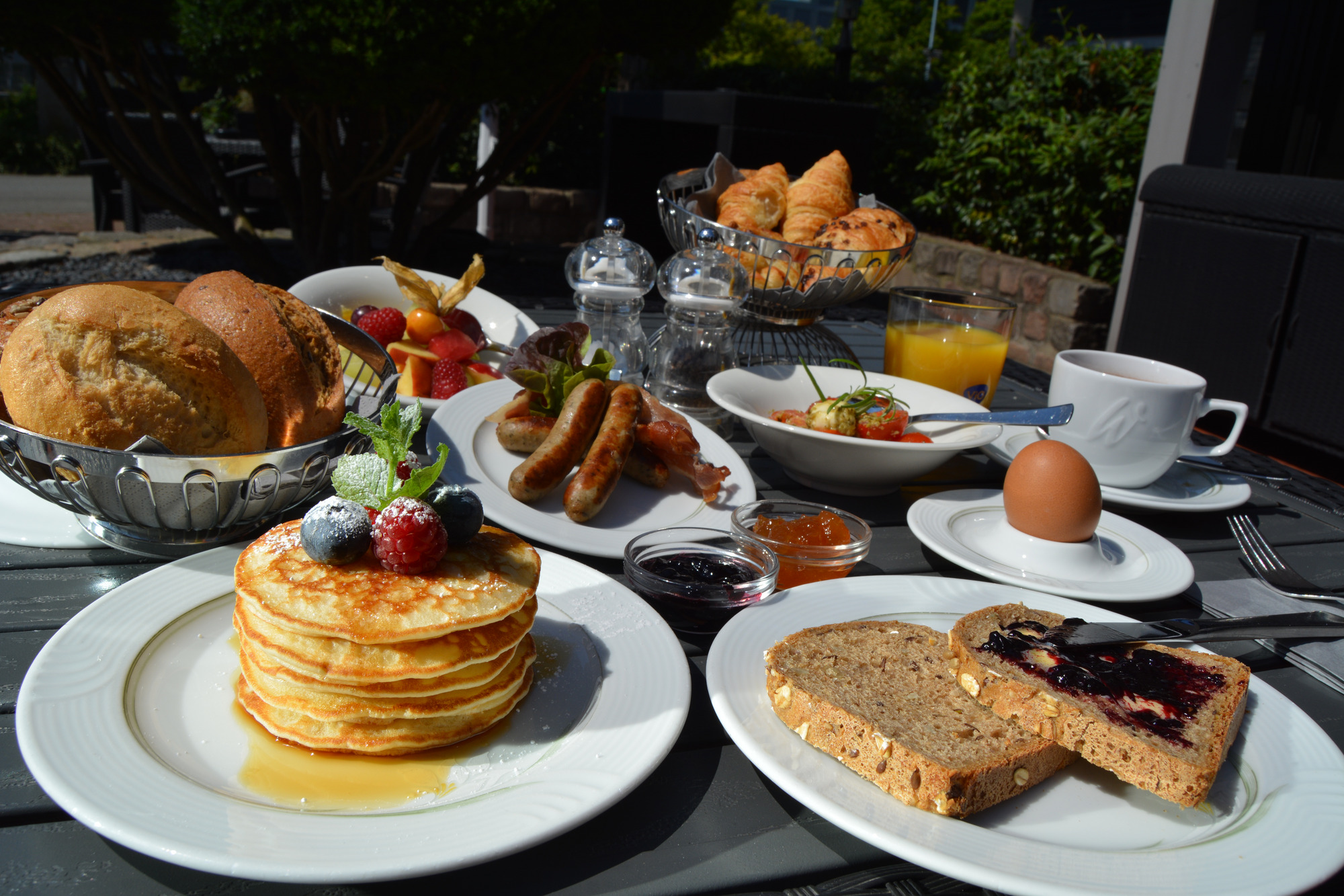 Frühstückstisch im Freien mit Gebäck, Brot, Obst, Ei, Orangensaft und Kaffee – Terrassenambiente.