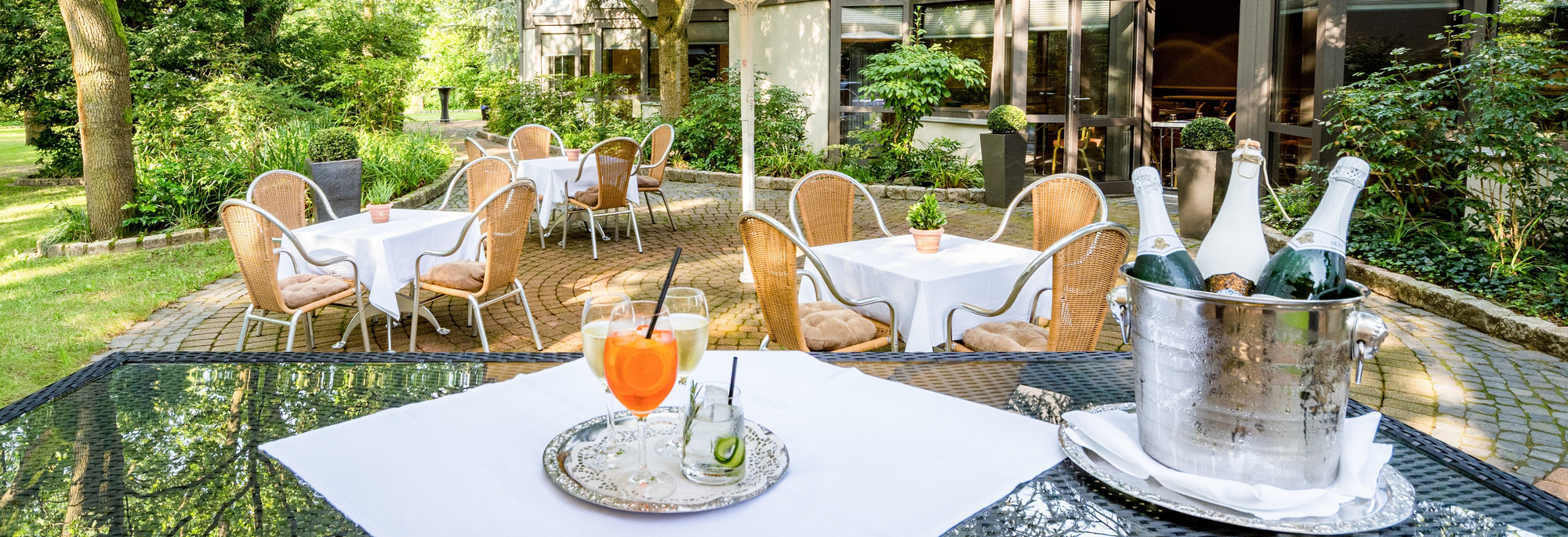 Gartenterrasse mit gedecktem Tisch unter gelbem Sonnenschirm, umgeben von Stühlen und grüner, ruhiger Naturkulisse.