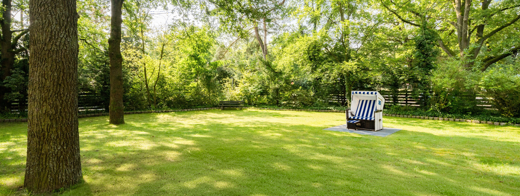 Grüner Park mit hohen Bäumen, gepflegtem Rasen, sonniger Atmosphäre und einem blau-weißen Strandkorb.