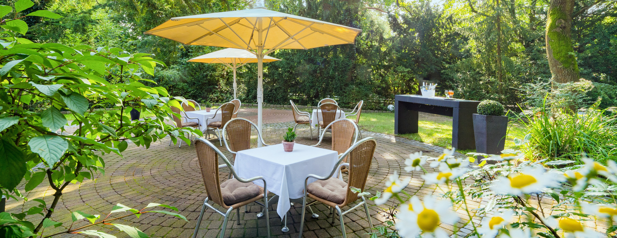 Garten mit Tisch, weißer Tischdecke, Holzstühlen, gelbem Sonnenschirm und blühender, grüner Umgebung.