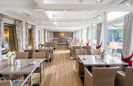 Eleganter Speisesaal mit weiß gedeckten Tischen, Blumenschmuck und viel Tageslicht durch große Fenster.