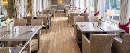 Eleganter Speisesaal mit weiß gedeckten Tischen, Blumenschmuck und viel Tageslicht durch große Fenster.