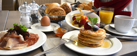 Frühstückstisch mit Pancakes, Eiern, Speck, Obstsalat, Saft und Kaffee – einladend und reich gedeckt.