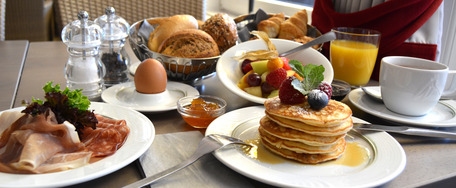 Frühstückstisch mit Pancakes, Eiern, Speck, Obstsalat, Saft und Kaffee – einladend und reich gedeckt.