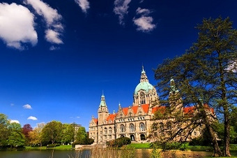 Neues Rathaus Hannover mit großer Kuppel und Türmen, gelegen an einem Teich, umgeben von Bäumen und Grün bei klarem, sonnigem Himmel.