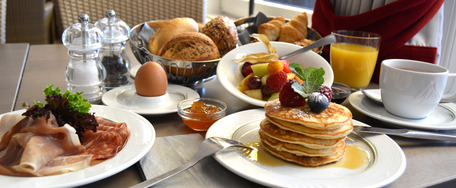 Frühstückstisch mit Pancakes, Eiern, Speck, Obstsalat, Saft und Kaffee – einladend und reich gedeckt.