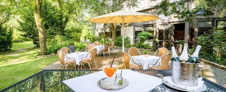 Gartenterrasse mit Tischen, weißen Tischdecken, orangefarbenen Stühlen, gelbem Sonnenschirm und grüner, ruhiger Umgebung.