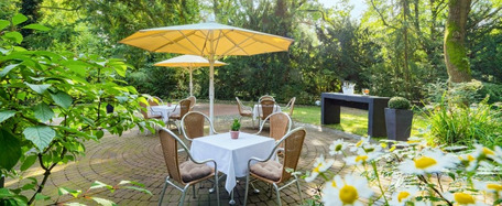 Garten mit Tisch, weißer Tischdecke, Holzstühlen, gelbem Sonnenschirm und blühender, grüner Umgebung.