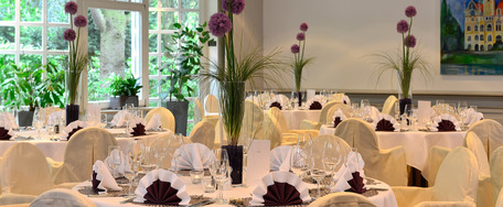 Eleganter Bankettsaal mit runden Tischen, weißen Tischdecken, Blumenschmuck, Stuhlhussen mit Goldschleifen und großen Fenstern.