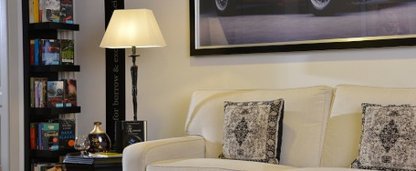 Gemütliche Wohnecke mit weißem Sofa, gemusterten Kissen, schwarzer Stehlampe und Bücherregal vor hellen Wänden und Holzfußboden.