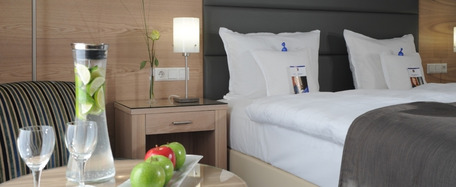 Hotelzimmer mit ordentlich gemachtem Bett, grauer Decke, Nachttisch mit Lampe und Tablett mit Wasser, Gläsern und Apfel im Vordergrund.