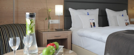 Hotelzimmer mit ordentlich gemachtem Bett, grauer Decke, Nachttisch mit Lampe und Tablett mit Wasser, Gläsern und Apfel im Vordergrund.