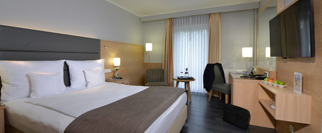 Modernes Hotelzimmer mit großem Bett, weißer Bettwäsche, Flachbild-TV, großen Fenstern und neutraler Deko.