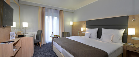 Modernes Hotelzimmer mit großem Bett, weißer Bettwäsche, Flachbild-TV, großen Fenstern und neutraler Deko.