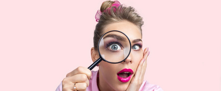 Person mit verschwommenem Gesicht, pinkem Shirt und Haaraccessoire, hält Lupe vor rosa Hintergrund.