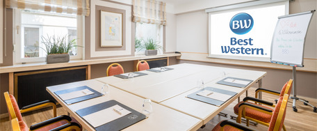 Konferenzraum mit rechteckigem Tisch (gelbe Tischdecke), acht Stühlen mit orangefarbenen Kissen, Notizmaterial, Wasser und Whiteboard.