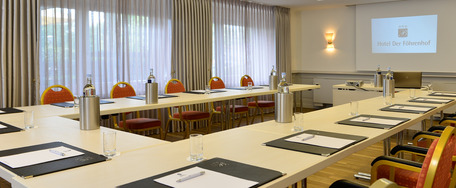 Konferenzraum mit langem Tisch, Stühlen, Notizblöcken, Projektorleinwand, Whiteboards und großen Fenstern.