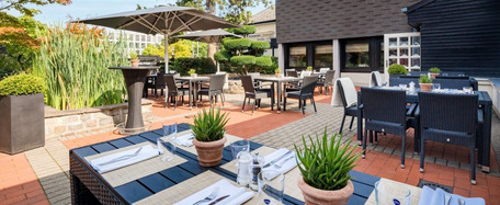 Außenterrasse mit Tischen, Stühlen, Sonnenschirm, Topfpflanzen und modernem Gebäude im Hintergrund bei sonnigem Wetter.